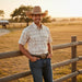 A man wearing a cowboy hat, a short-sleeve button-up shirt with a small black-on-cream pattern, and blue jeans stands outdoors in a rural setting with a wooden fence and field behind him.