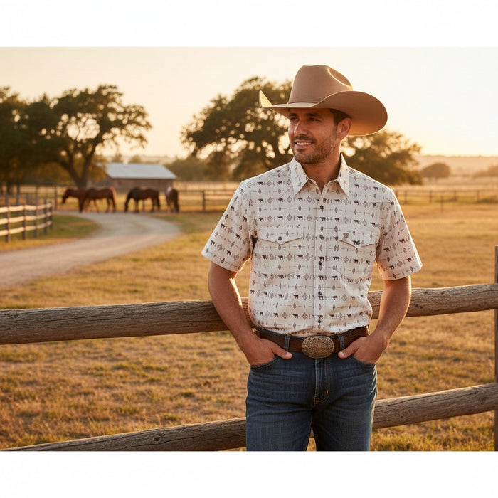 A man wearing a short-sleeve western button-up shirt with a small brown cowboy motif on a cream background, blue jeans, and a tan cowboy hat, standing in front of a wooden fence in a rural outdoor setting at sunset.
