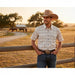 A man wearing a short-sleeve western button-up shirt with a small brown cowboy motif on a cream background, blue jeans, and a tan cowboy hat, standing in front of a wooden fence in a rural outdoor setting at sunset.