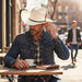 Man wearing a cowboy hat and denim jacket sitting at an outdoor cafe table.