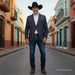A man wearing a dark navy blazer with contrasting shoulder patches, a white dress shirt, blue jeans, brown boots, and a black cowboy hat stands on a cobblestone street in a colorful town square.