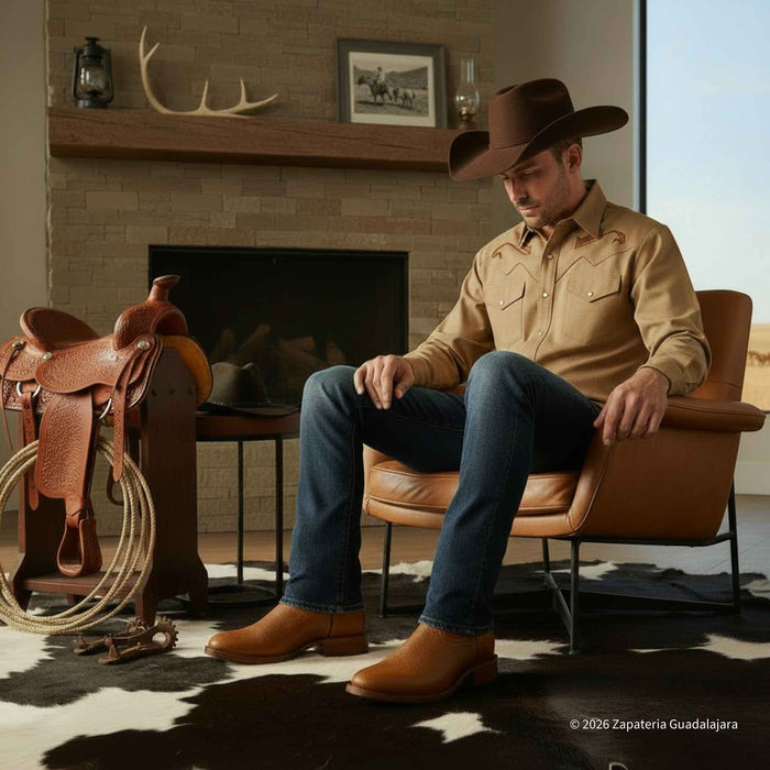 Man in cowboy attire sitting in a chair with a fireplace and saddle in the background