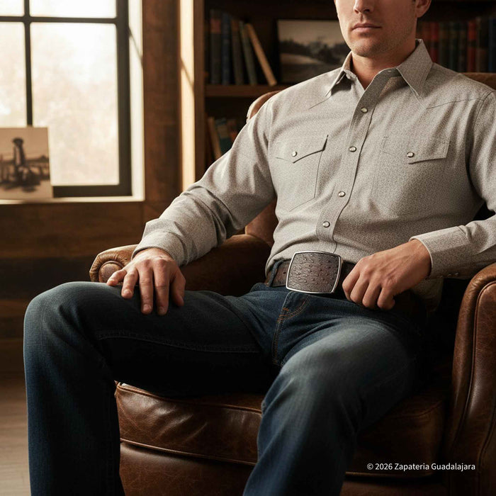 A man wearing a light beige western-style button-down shirt and dark blue jeans sits in a brown leather armchair in a rustic room with bookshelves and a window.