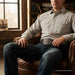 A man wearing a light beige western-style button-down shirt and dark blue jeans sits in a brown leather armchair in a rustic room with bookshelves and a window.