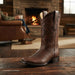 A brown leather cowboy boot with decorative stitching stands on a rustic wooden table in a cozy living room setting with a fireplace and a person sitting in an armchair in the background.