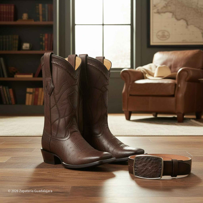 A pair of dark brown leather cowboy boots with decorative stitching, displayed on a wooden floor next to a matching brown belt in a rustic living room setting.
