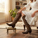 Woman in a white dress wearing brown cowboy boots in a cozy living room.