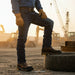 Person wearing work boots and gloves on a construction site with machinery in the background.