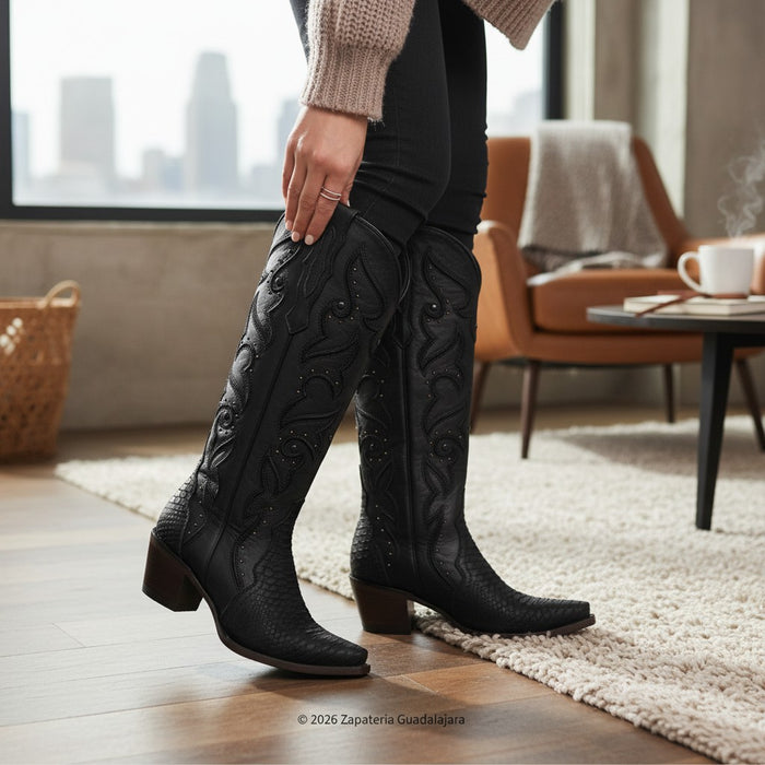 A person wearing black knee-high western boots with carved floral detailing and stacked block heels, standing on a light rug in a modern living room. The boots are the clear product focus, with a city view through a window in the background.