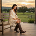 A woman sits on a wooden porch bench wearing a cream lace dress and dark brown cowboy boots, with a rural landscape at sunset in the background.