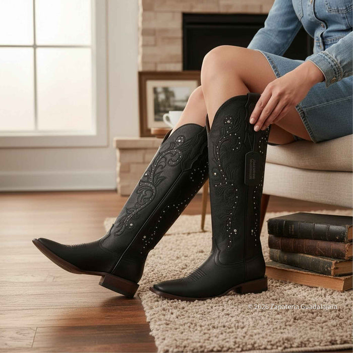 Person wearing black cowboy boots with intricate designs, sitting on a couch in a cozy living room.