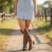 Person wearing a denim dress and brown cowboy boots on a dirt path with blurred background