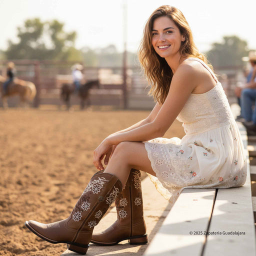 Woman sitting on a wooden bench wearing brown embroidered boots and a white dress in an outdoor setting with horses in the background.