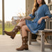 Woman in a blue dress and brown cowboy boots sitting on a wooden bench outdoors.