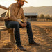 A man in a tan long-sleeve western shirt, dark blue jeans, and brown boots sits on a wooden bench in a sunlit outdoor ranch setting with mountains and a barn in the background.