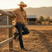 A man in a tan cowboy hat, tan long-sleeve button-up shirt, dark blue jeans, and brown boots leans against a wooden fence in a sunlit ranch paddock with mountains in the background.