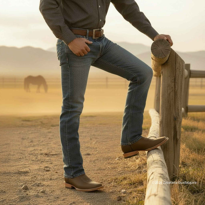 A man wearing blue jeans, a dark gray long-sleeve shirt, and brown leather boots leans on a wooden fence in a dusty outdoor ranch setting with mountains and a horse in the background.