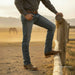 A man wearing blue jeans, a dark gray long-sleeve shirt, and brown leather boots leans on a wooden fence in a dusty outdoor ranch setting with mountains and a horse in the background.