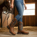 Person wearing brown cowboy boots with intricate designs, standing next to a horse in a stable.