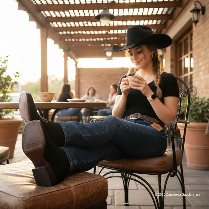 Woman sitting outdoors on a patio, wearing a cowboy hat and boots, using a smartphone.